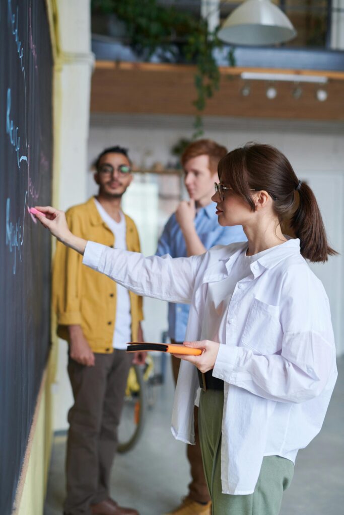 pexels-photo-3184644-3184644-1 A group of young people discussing ideas at a blackboard in a modern office setting.